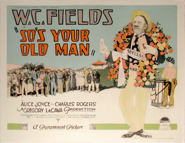 W.C. Fields wearing a garland of roses around his neck while holding-up a golf ball.